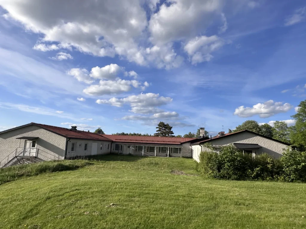Låg tegelbyggnad med rött tak och grön gräsmatta under en blå sommarhimmel.